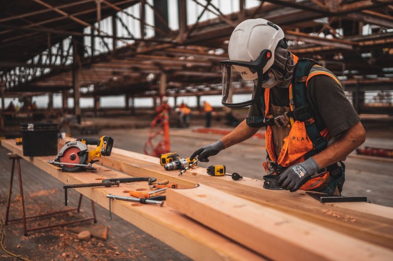 A student waring a hardhat and sheild visor works on a long slab of lumber with drilling and clamping tools strewn about them