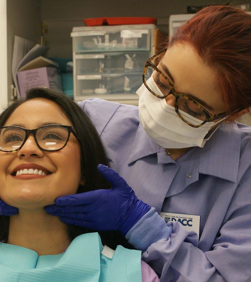A DACC dentist in training, wearing the appropriate attire and facemask, carefully examines the jowls of her smiling patient.